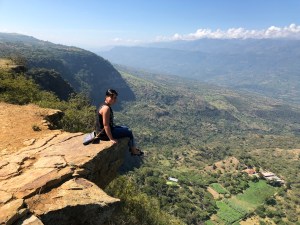 Catalina sitting on the edge of a cliff at El Salto del Mico in Barichara, Colombia, overlooking a vast green valley and distant mountains.