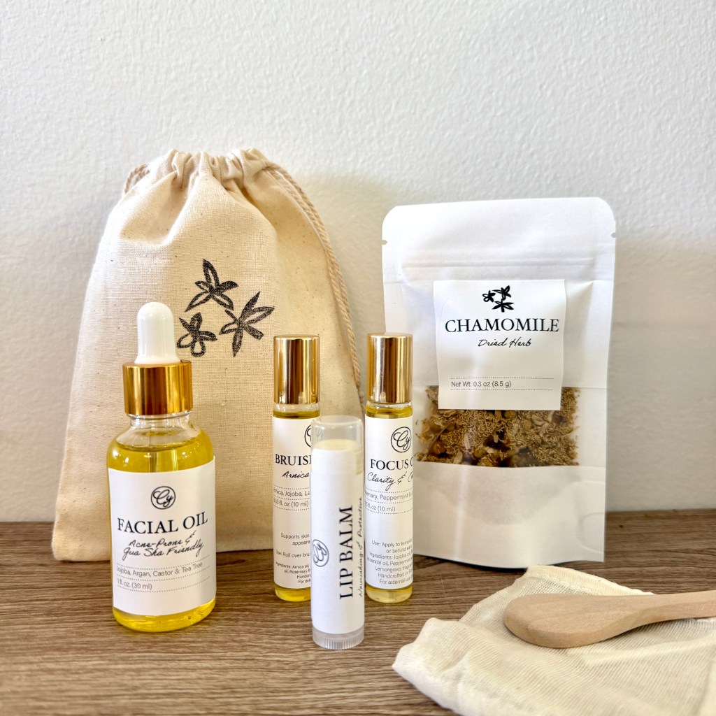 A minimalist photo of handmade herbal skincare products including facial oil, roller blends, lip balm, dried chamomile, and a cotton pouch arranged on a wooden surface in soft natural light.